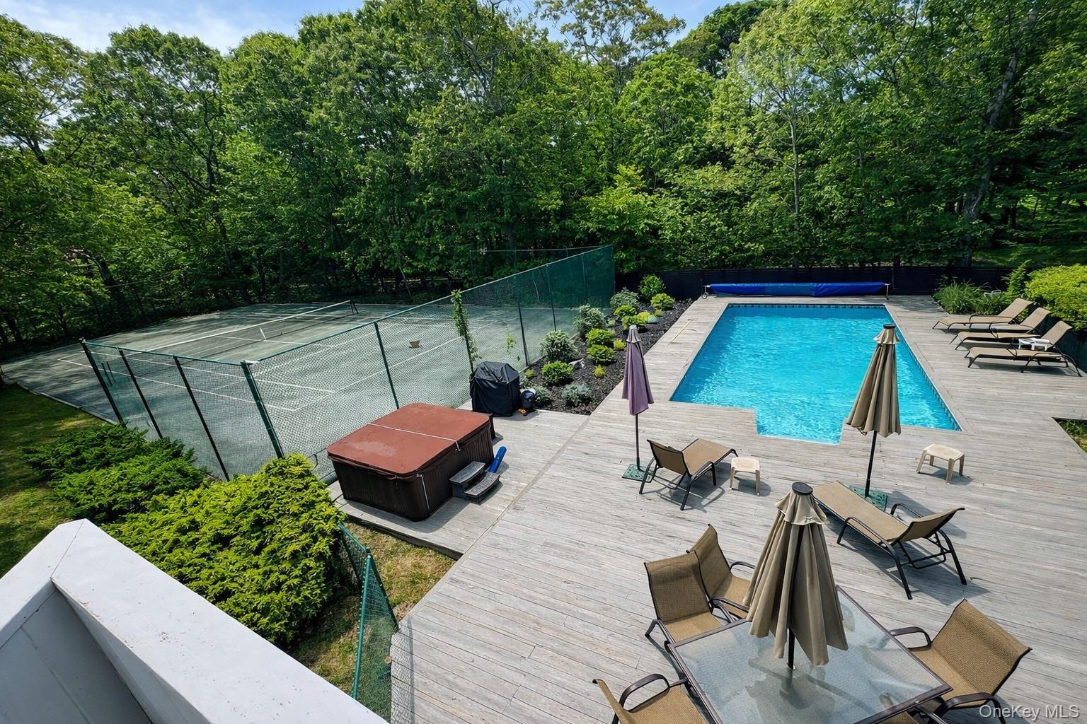 Swimming pool and broad wood deck beside a fenced tennis court, hot tub, and wooded surroundings.