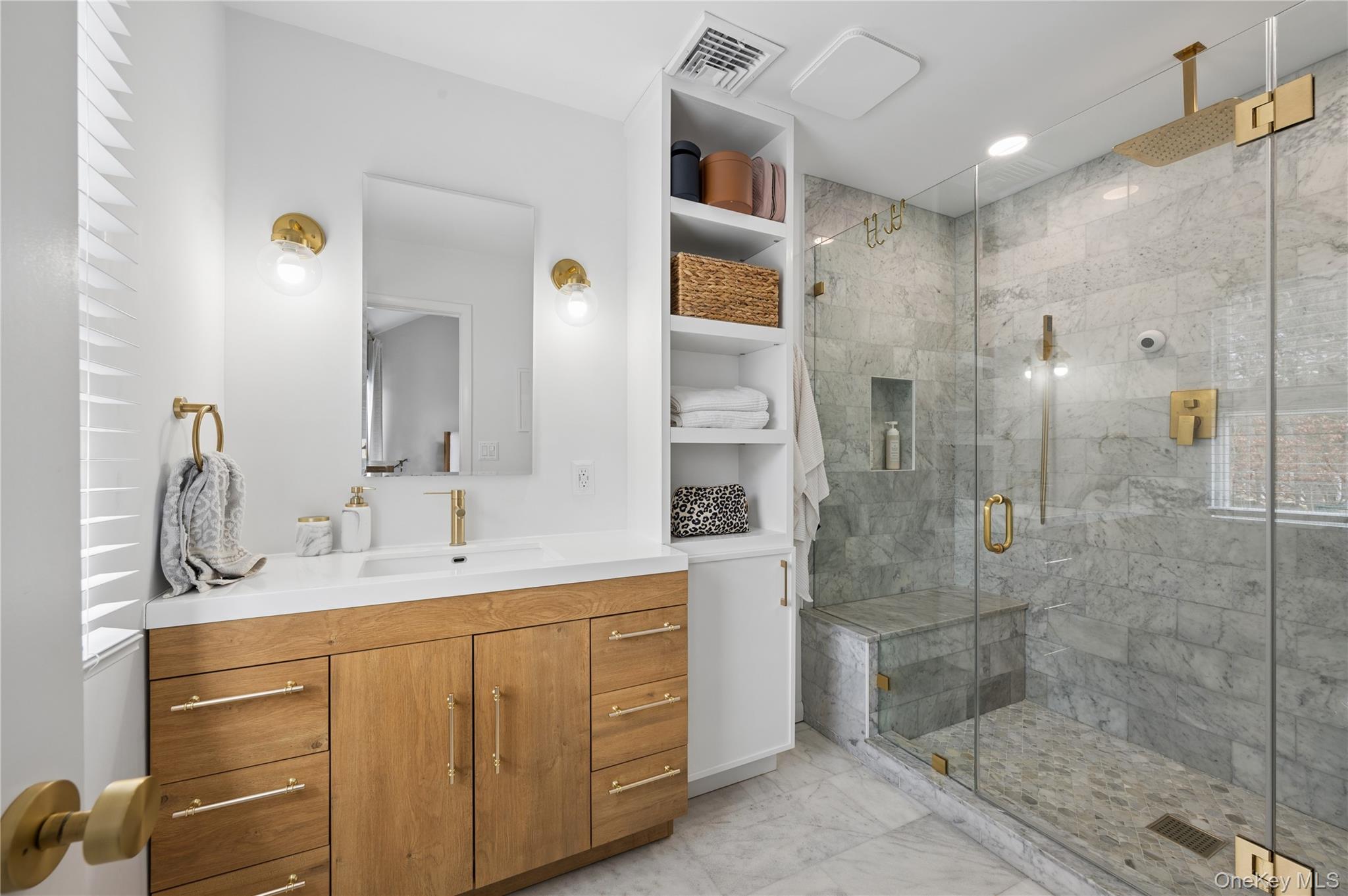 Bathroom with a wood vanity and brass fixtures beside a glass-enclosed marble shower with a bench.