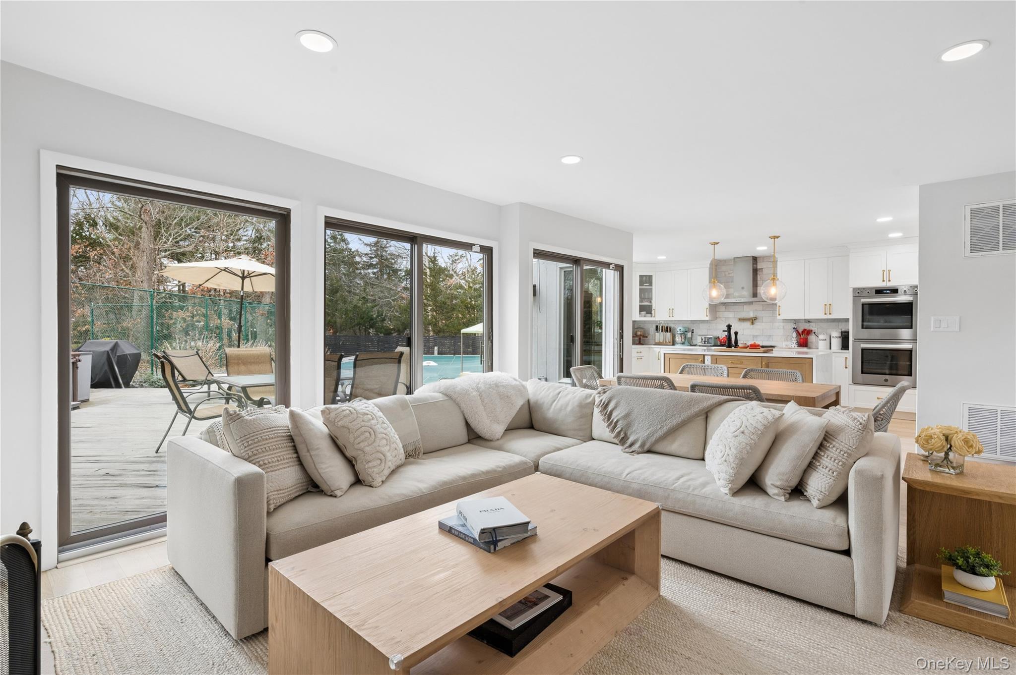 Open-plan living room with a large sectional sofa, white kitchen, and sliding doors overlooking a pool.