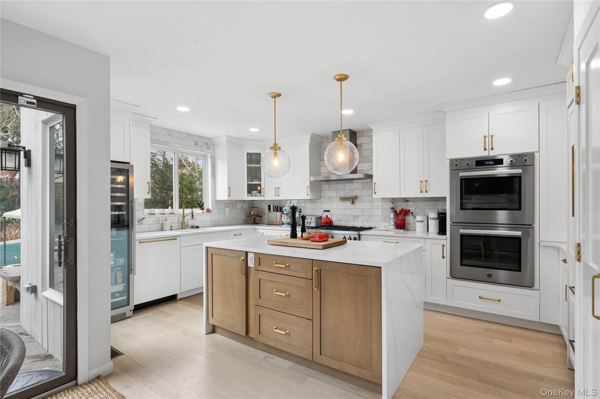 Bright white kitchen with a wood-paneled island, brass accents, and stainless steel double ovens.