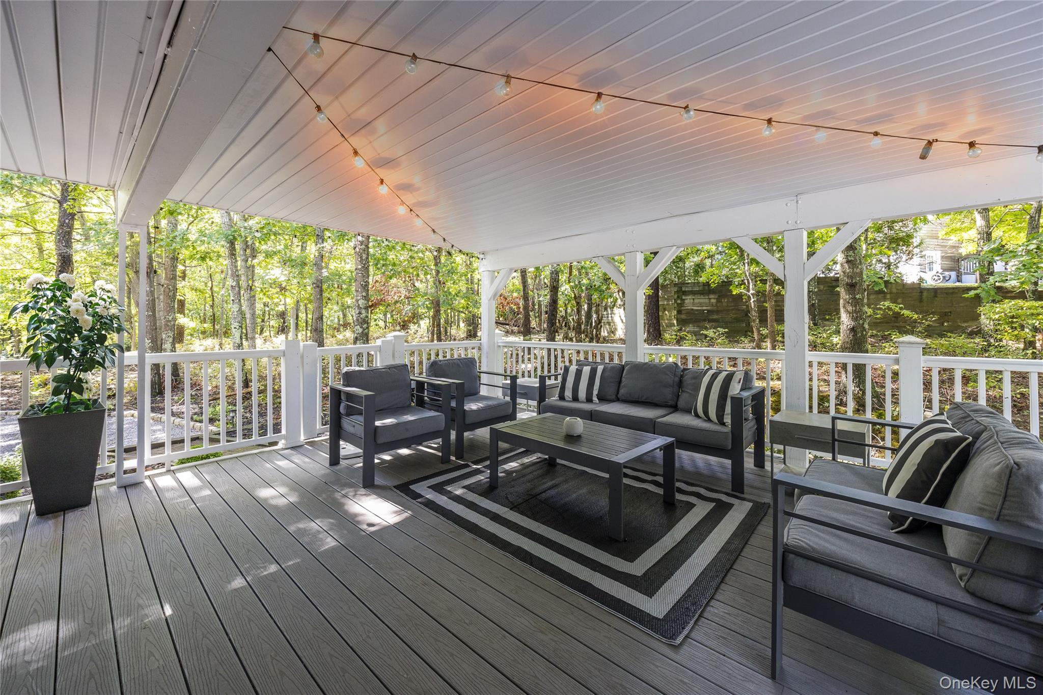 Covered deck furnished with gray lounge seating and string lights, surrounded by leafy trees.