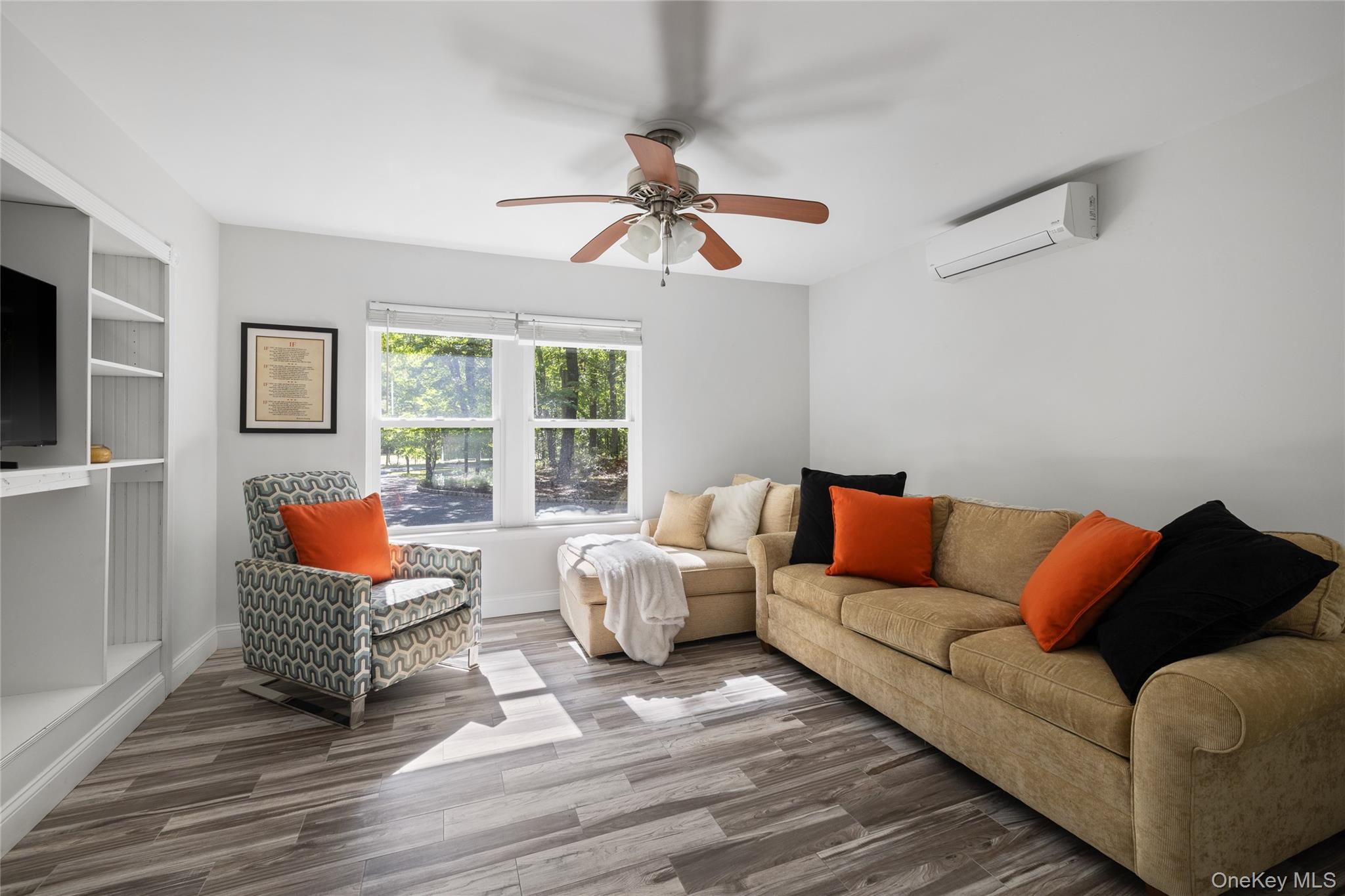 Living room with a tan sofa, orange accent pillows, patterned armchair, and windows overlooking trees.