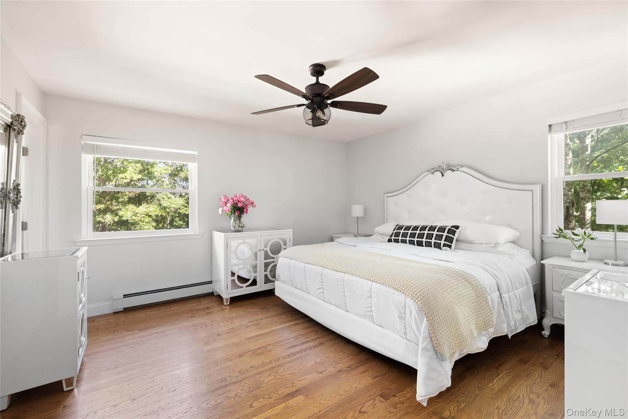 Staged bedroom with a white upholstered bed, hardwood floors, and leafy views through two windows.