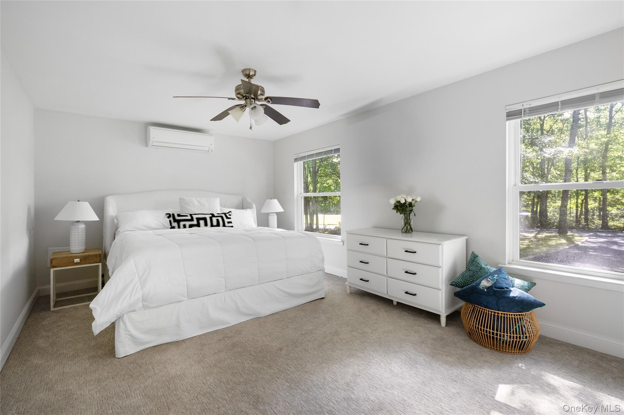 Staged bedroom with a white bed, dresser, and windows overlooking trees.