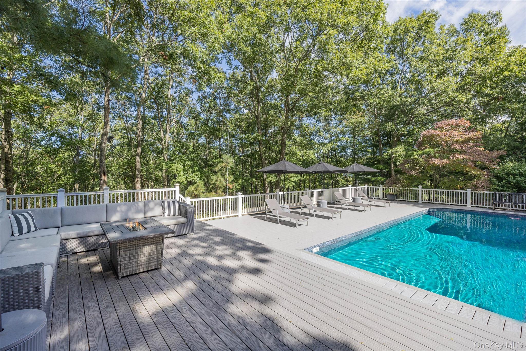 Backyard swimming pool beside a furnished gray deck, lounge chairs, and mature trees.