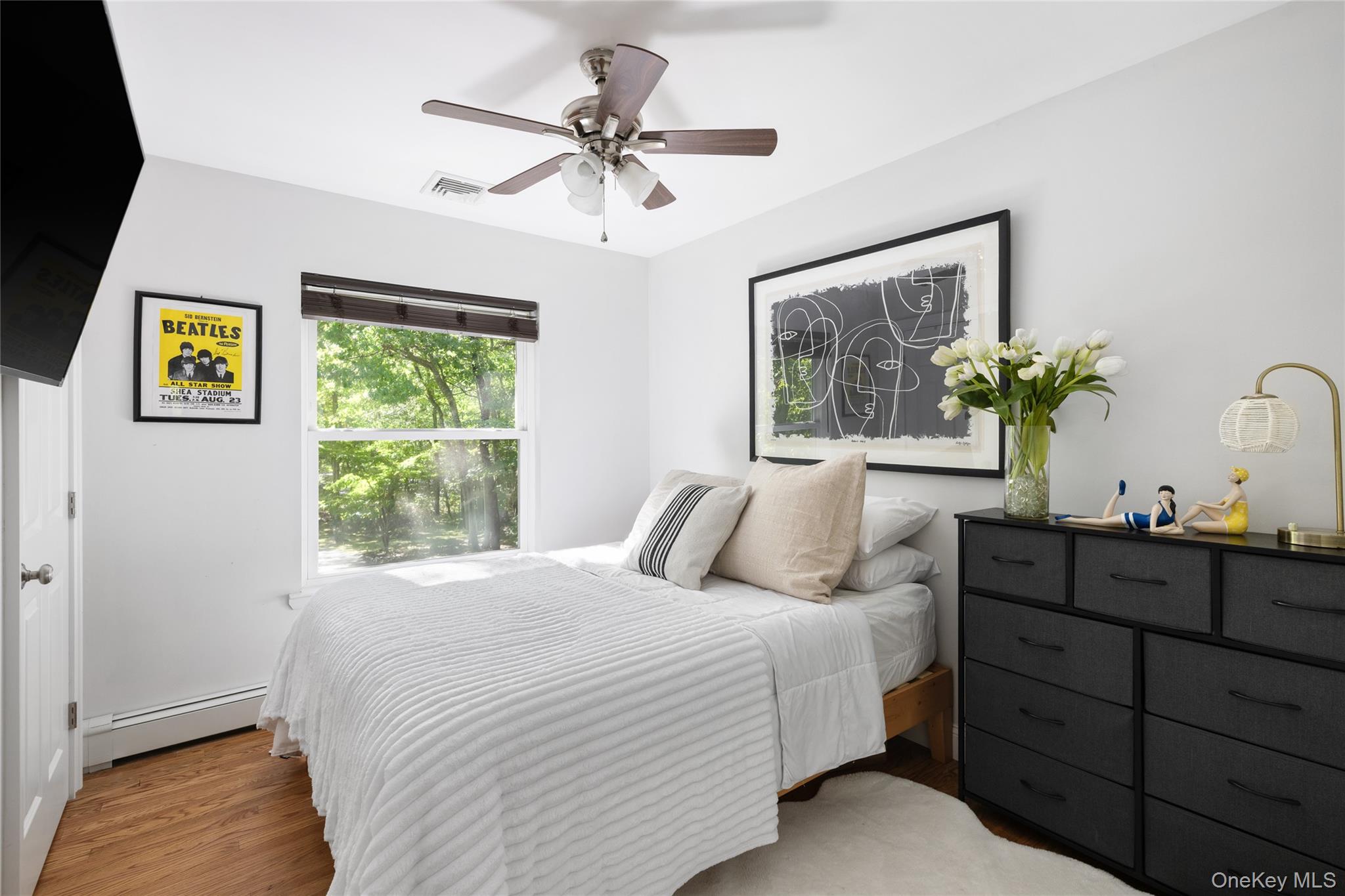 Bedroom with a white quilted bed, dark dresser, and window overlooking leafy trees.