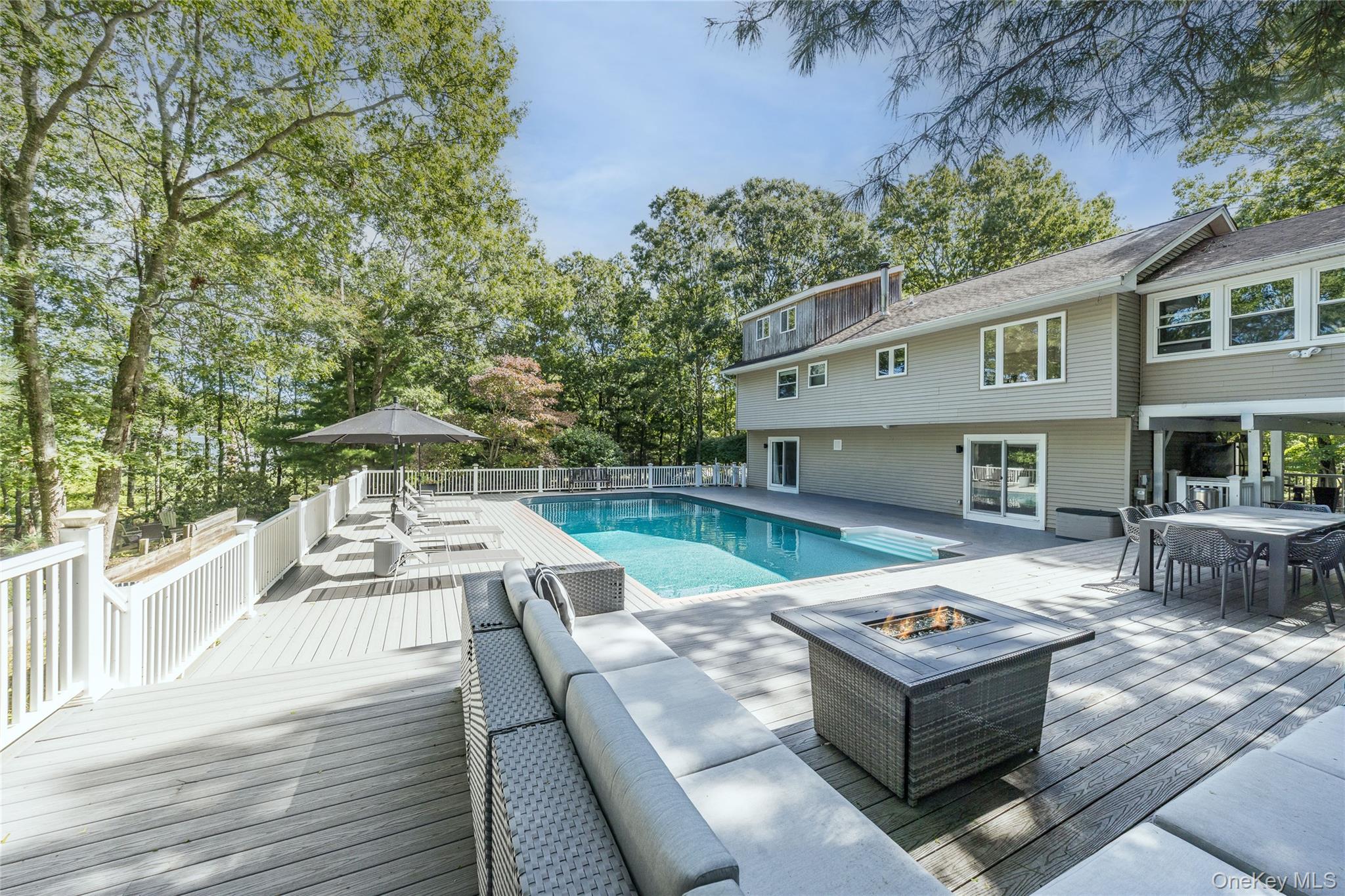Home’s rear deck with swimming pool, lounge seating, and fire table among mature trees.