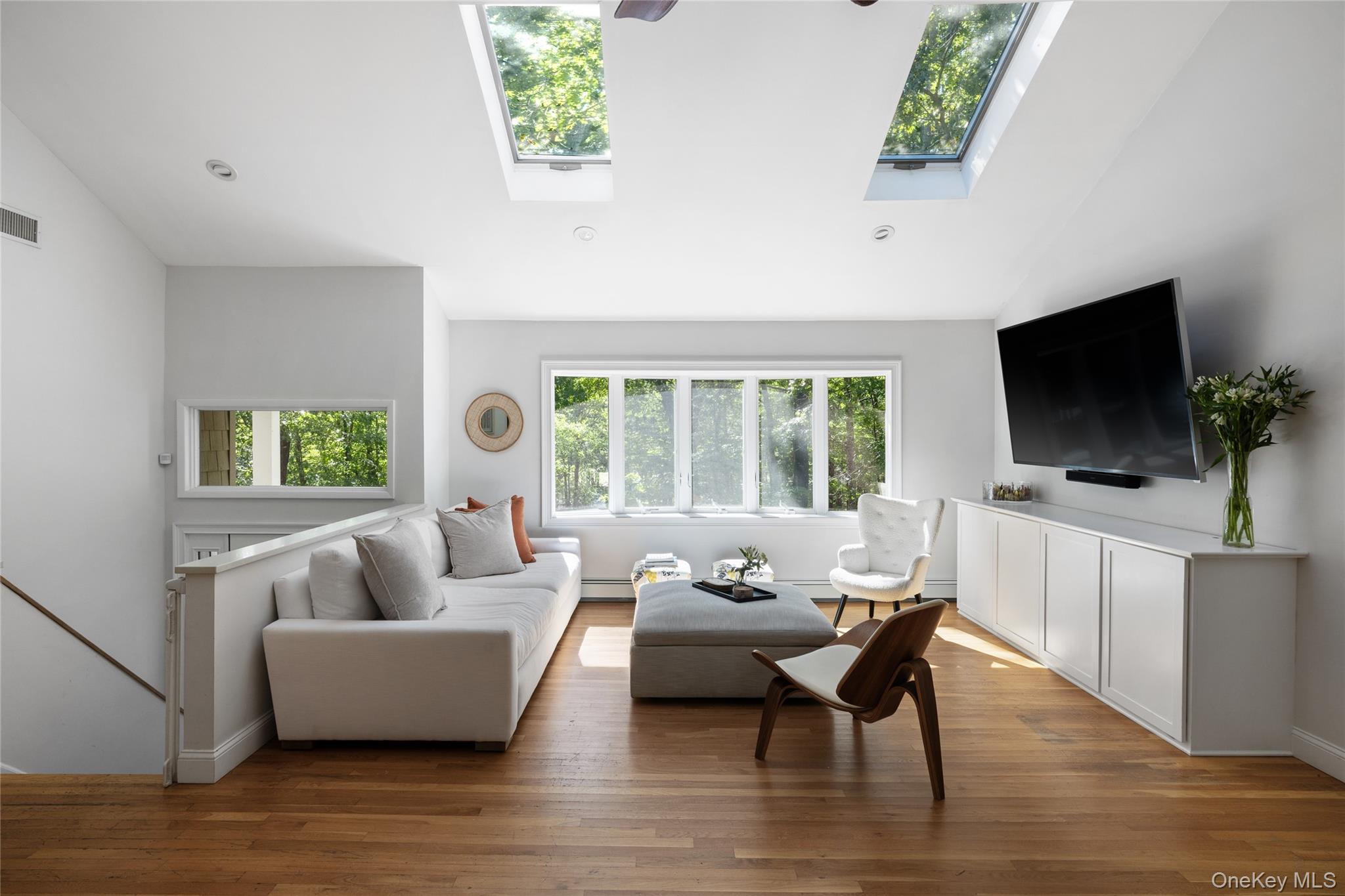 Bright living room with skylights, hardwood floors, and wide windows overlooking trees.
