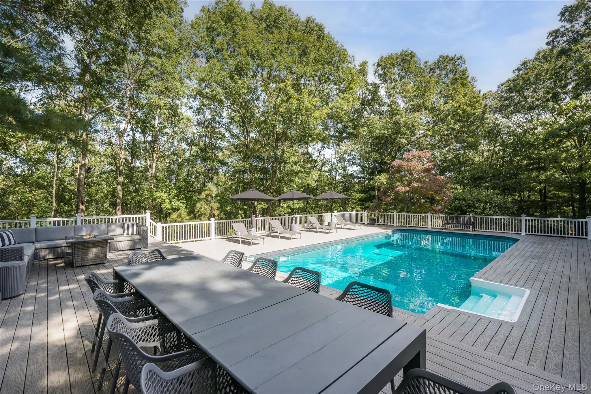 Backyard pool with a gray deck, outdoor dining area, lounge chairs, and mature trees.