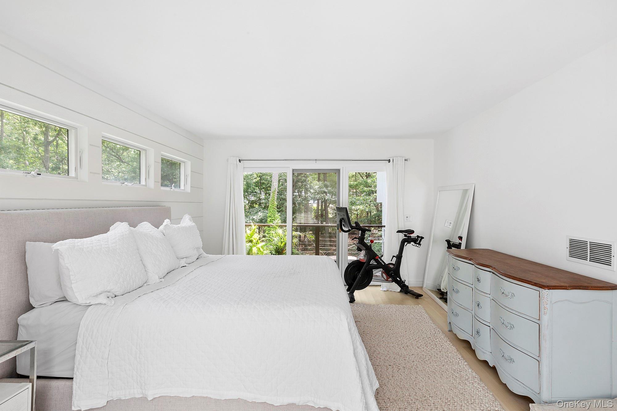 Bright bedroom with a white bed, pale blue dresser, and exercise bike by wooded views.