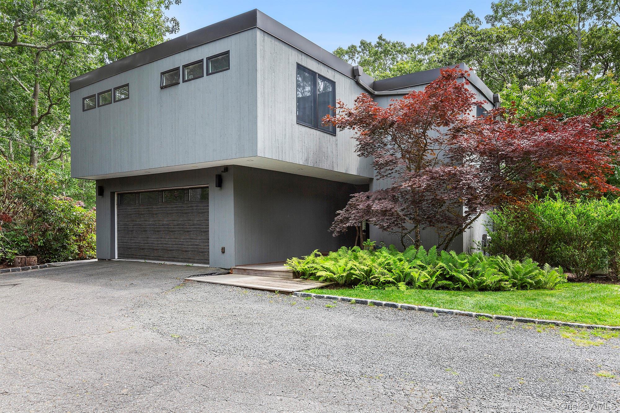 Modern gray two-story home with a flat roof, garage, gravel driveway, and landscaped entrance.