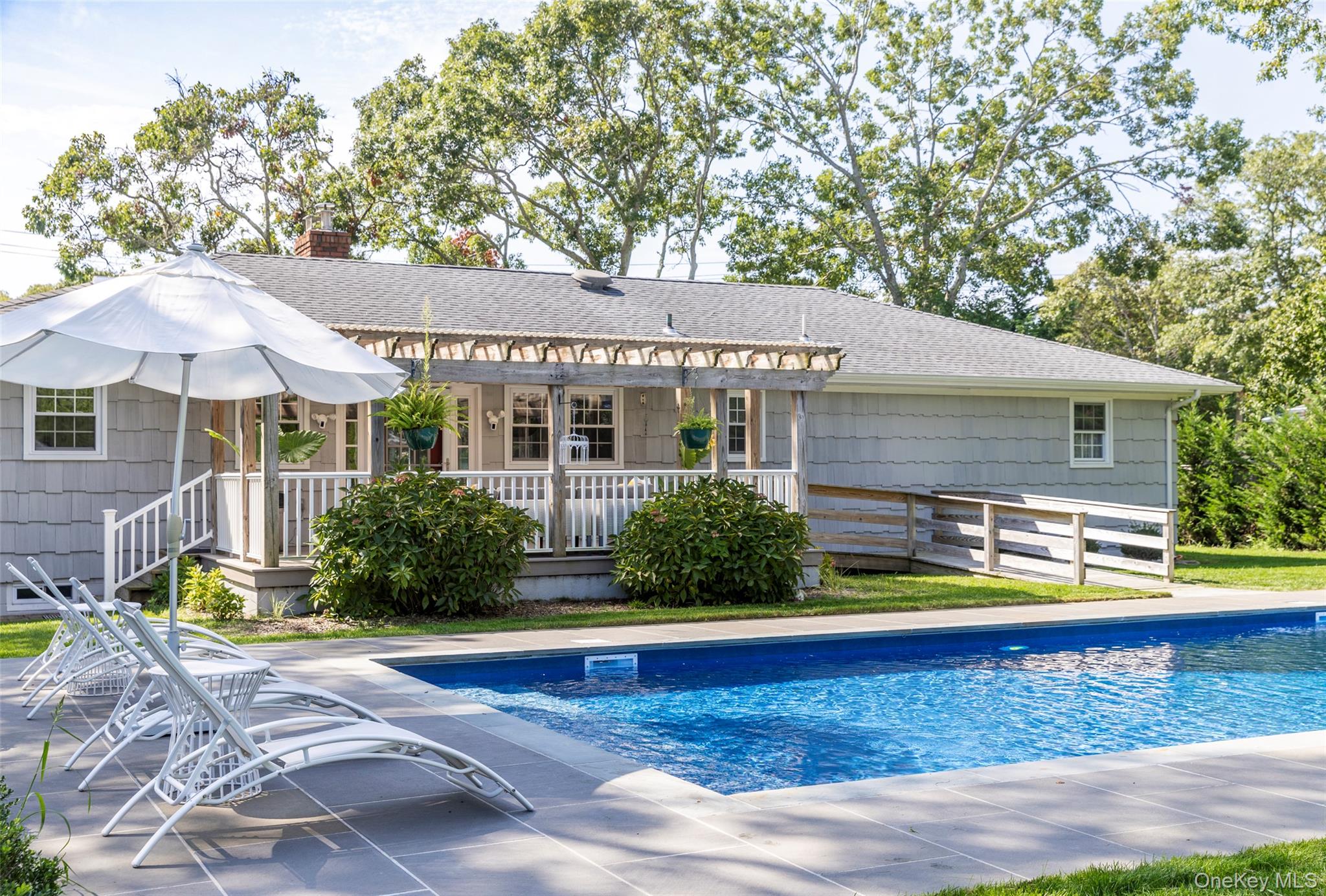 Gray shingle-sided home with a covered porch and ramp beside a swimming pool and white lounge chairs.