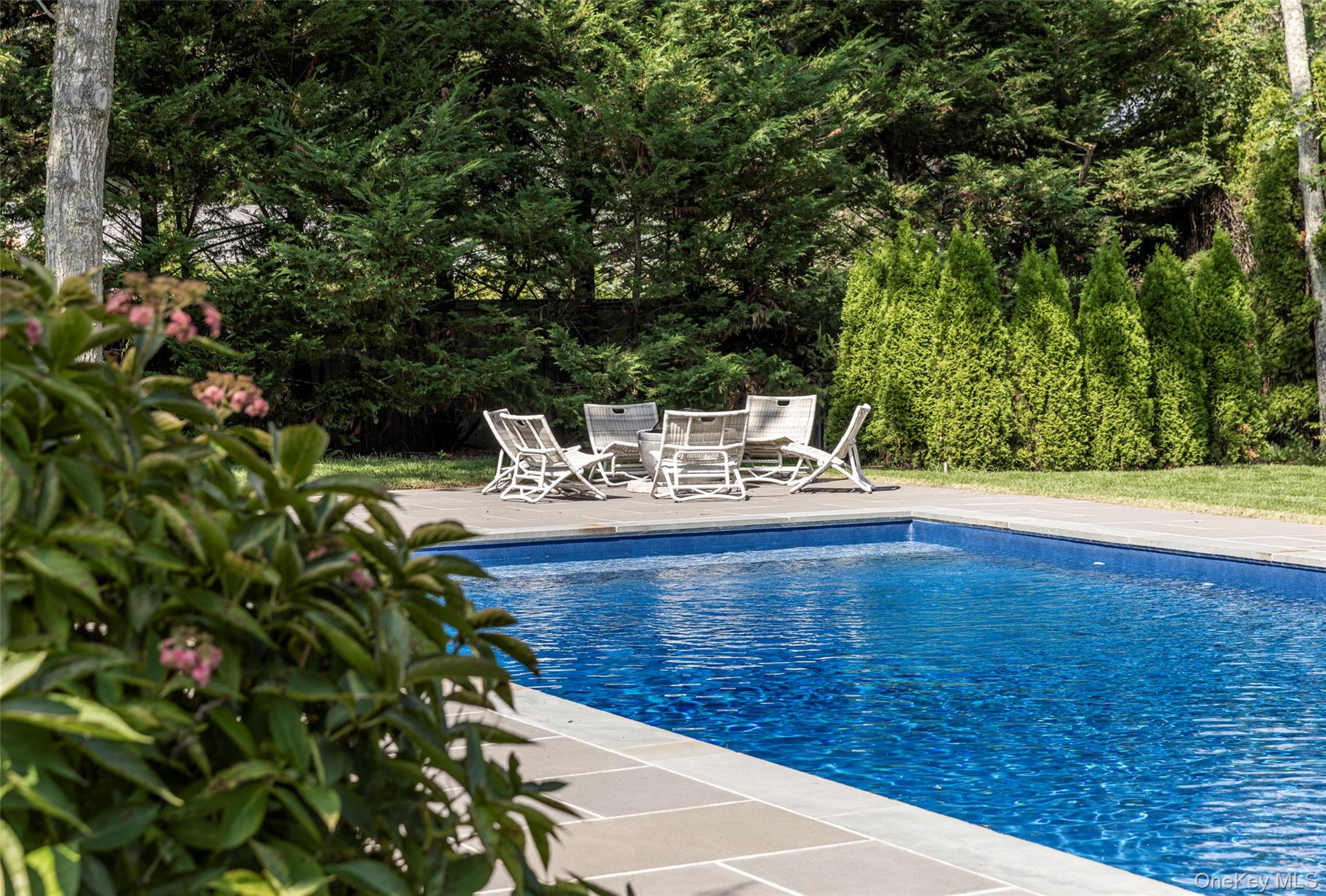 Outdoor swimming pool beside a stone patio with white lounge chairs and tall evergreen hedges.