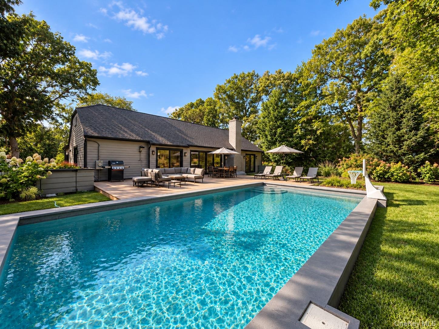 Modern gray home behind a swimming pool, with a furnished patio and mature trees.
