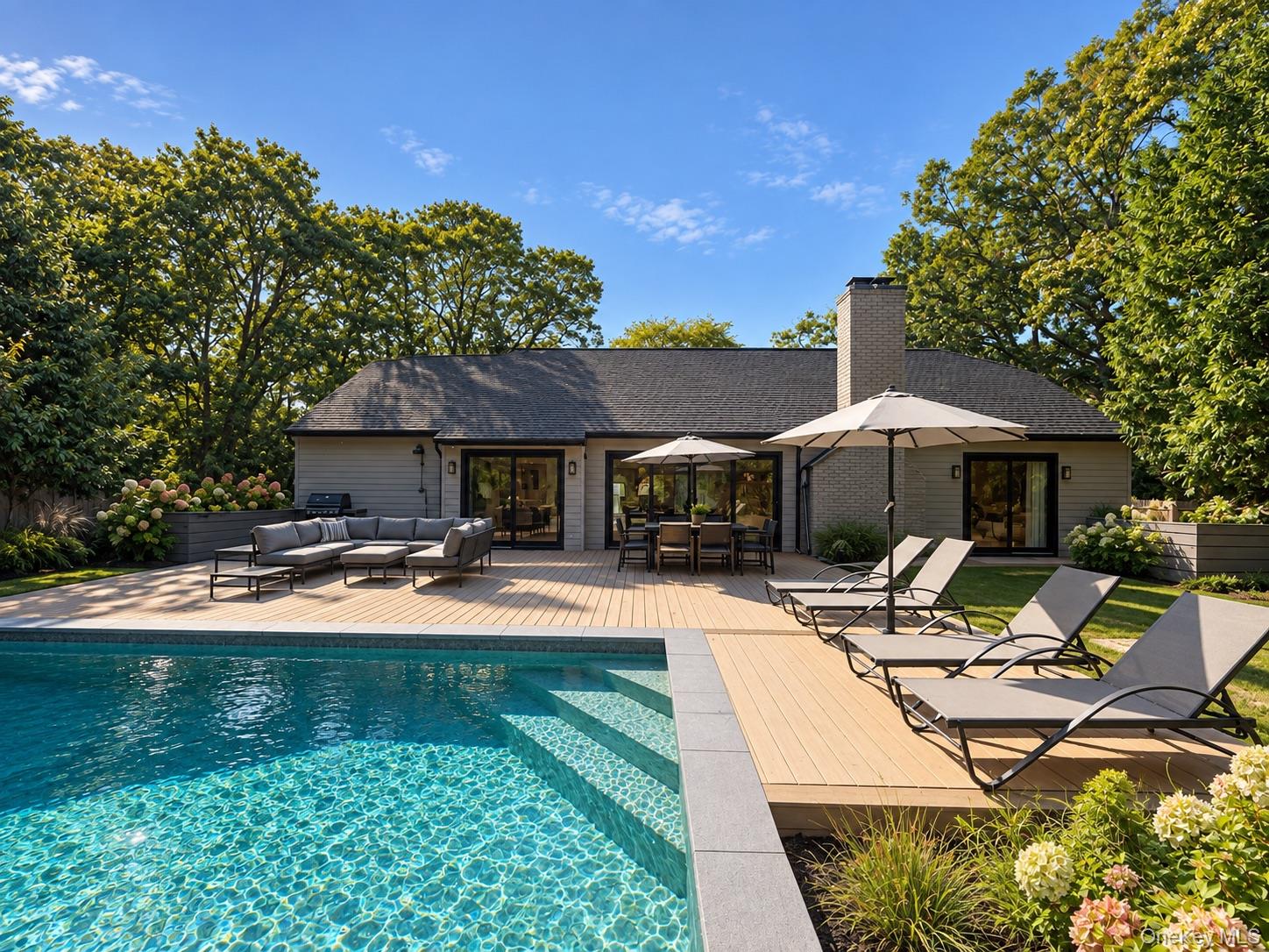 Single-story gray home behind a swimming pool and furnished deck with umbrellas and poolside loungers.
