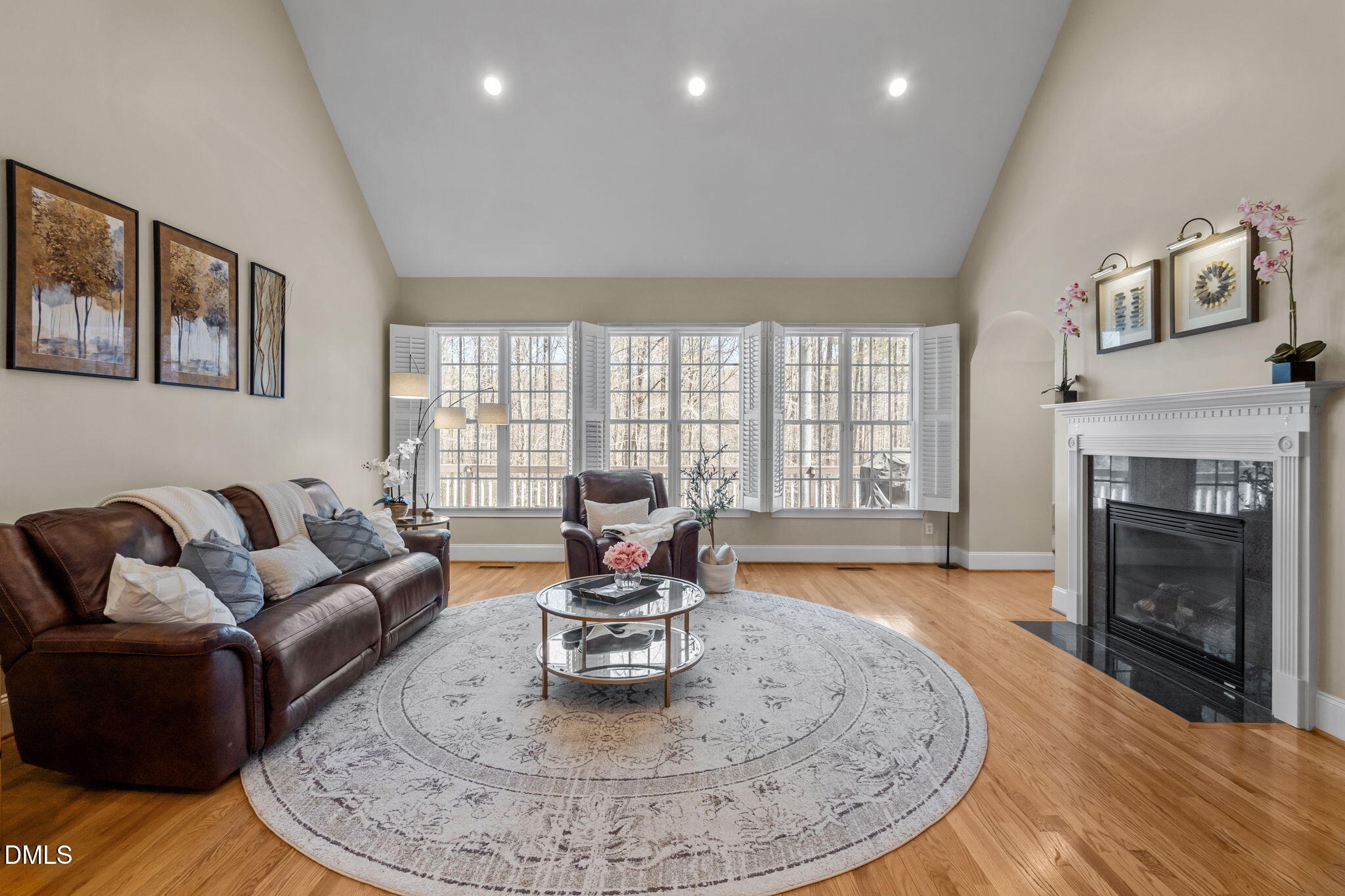 Living room with a leather sofa, white fireplace, round rug, and three windows overlooking leafless trees.