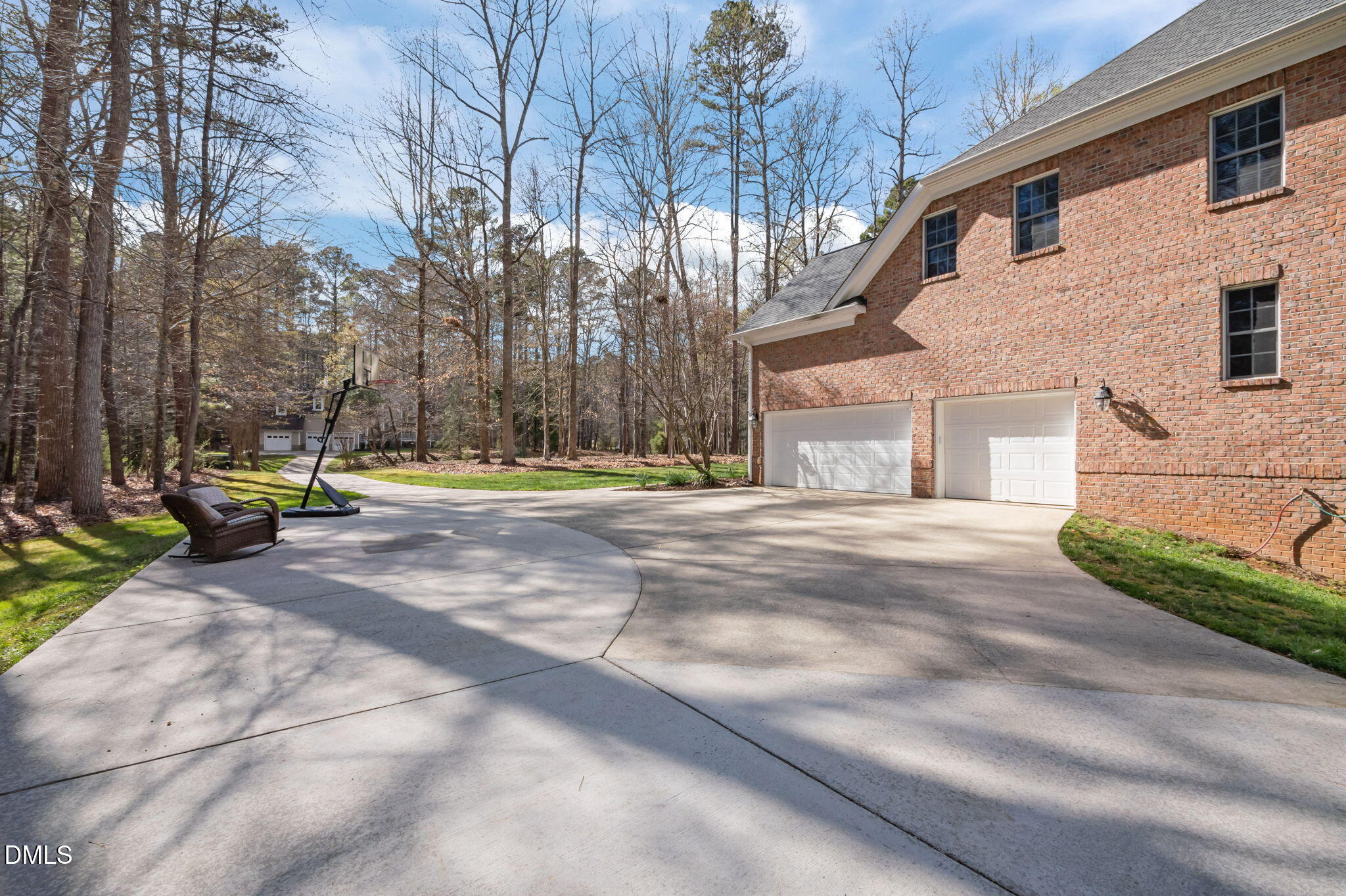 Brick house with two garage doors beside a curved driveway, basketball hoop, and wooded yard.