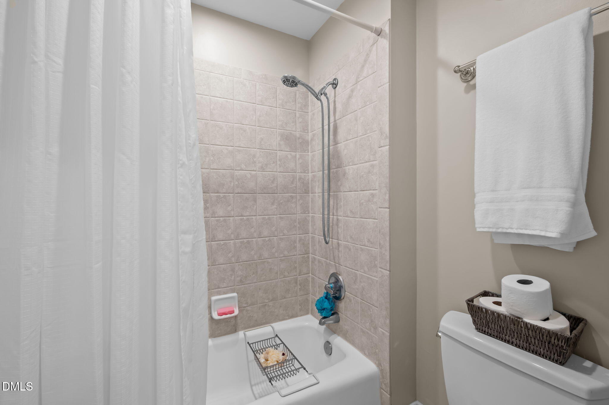 Bathroom with a tiled tub and shower, white curtain, handheld showerhead, and towels above the toilet.