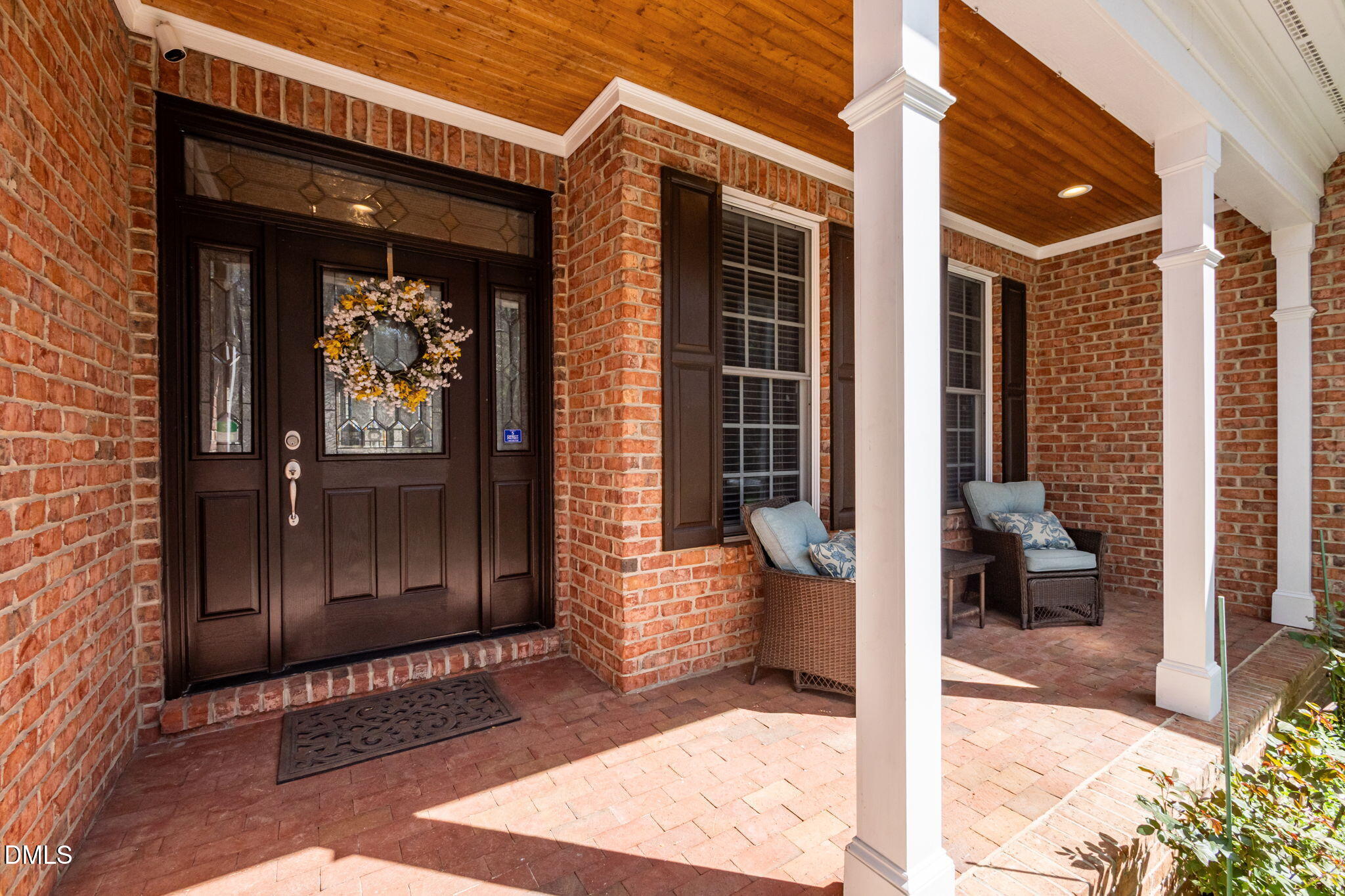 Brick front porch with a dark wood entry door, floral wreath, white columns, and wicker seating.