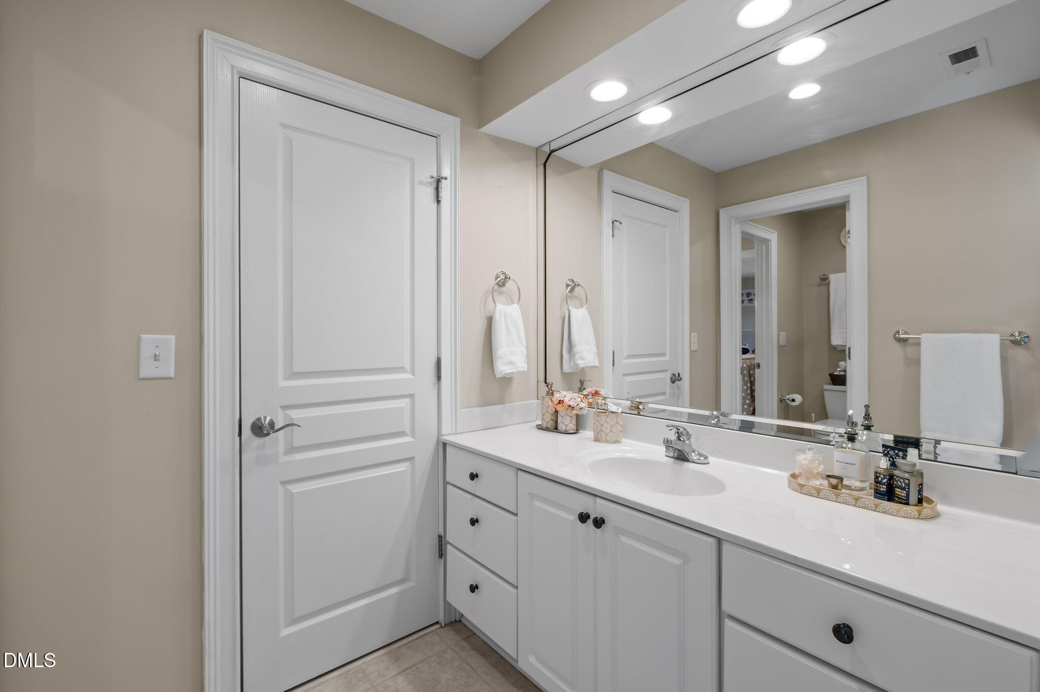 Bathroom with a white vanity and large mirror, viewed toward two doors and an adjoining room.
