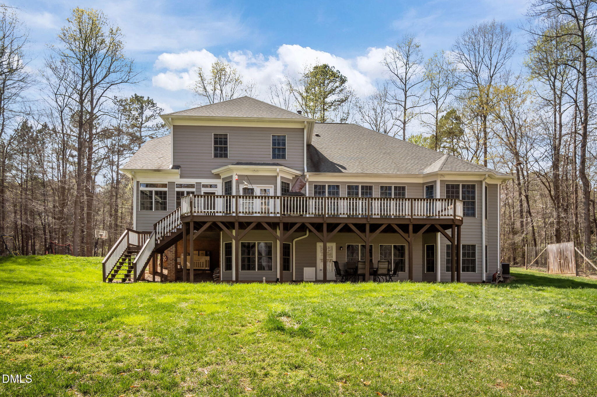Two-story gray house with a wide raised deck and covered patio overlooking a grassy, wooded yard.
