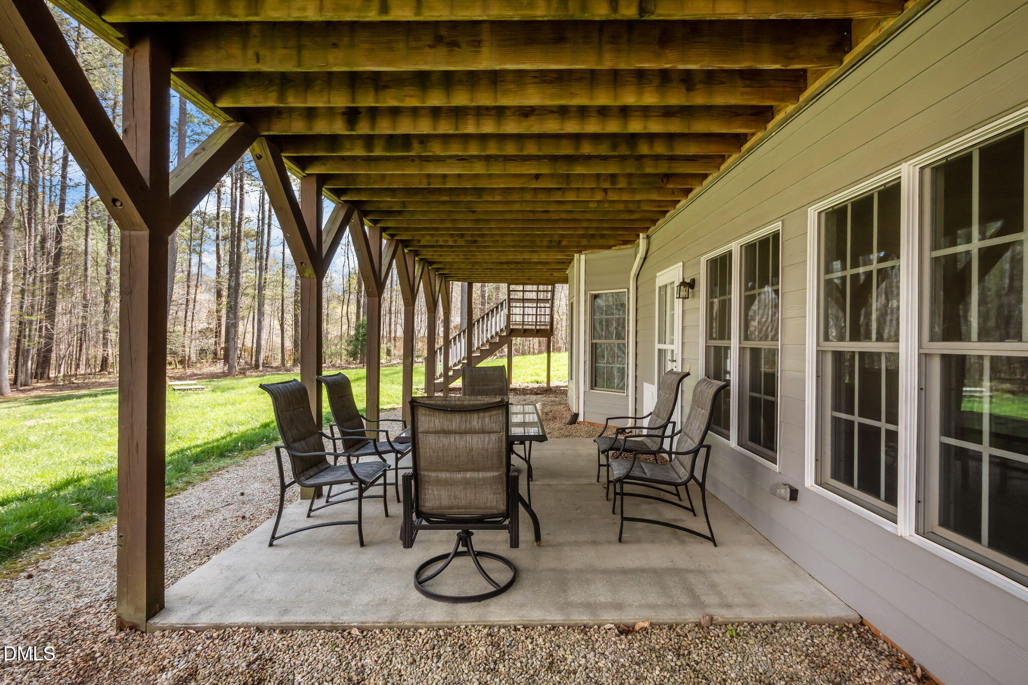 Covered patio with outdoor dining furniture, timber beams, and wooded lawn beyond.