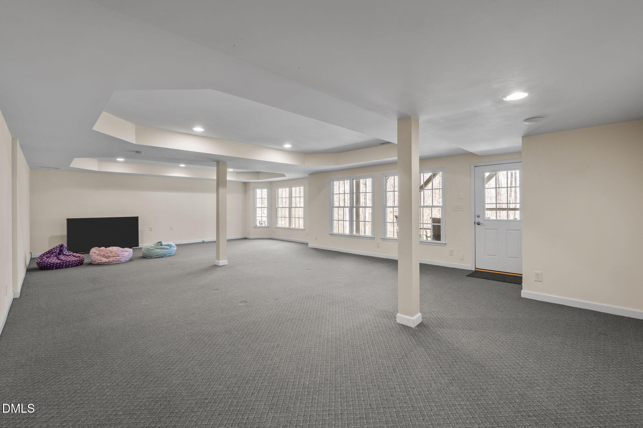 Finished basement with gray carpet, support columns, large windows, and a TV beside beanbag chairs.