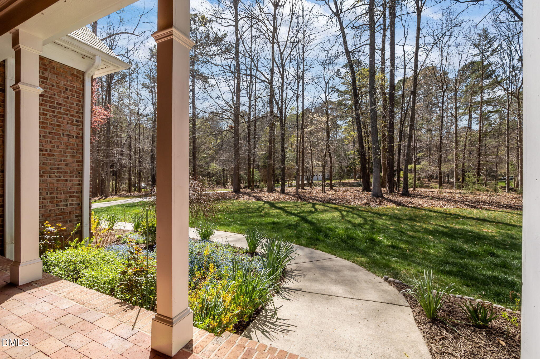 Brick porch with pale columns overlooking a curved walkway, landscaped lawn, and wooded yard.