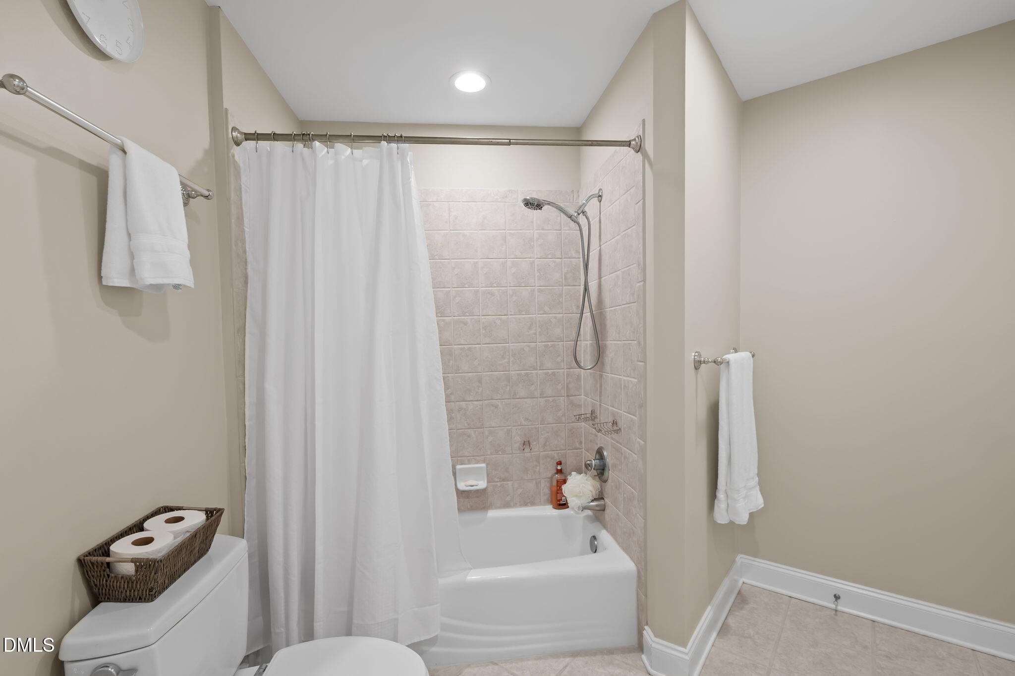 Bathroom with tiled tub and shower, white curtain, toilet, and chrome towel bars.