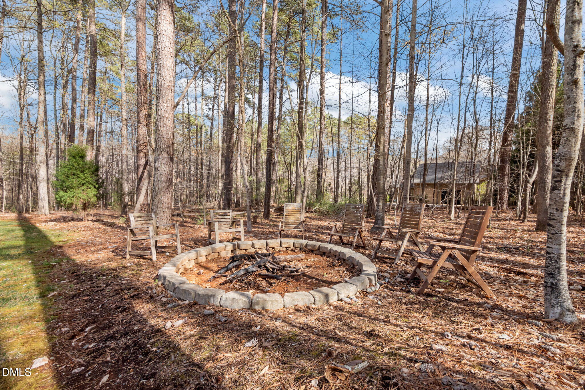 Wooded backyard fire pit ringed by wooden chairs, with a small outbuilding in the background.