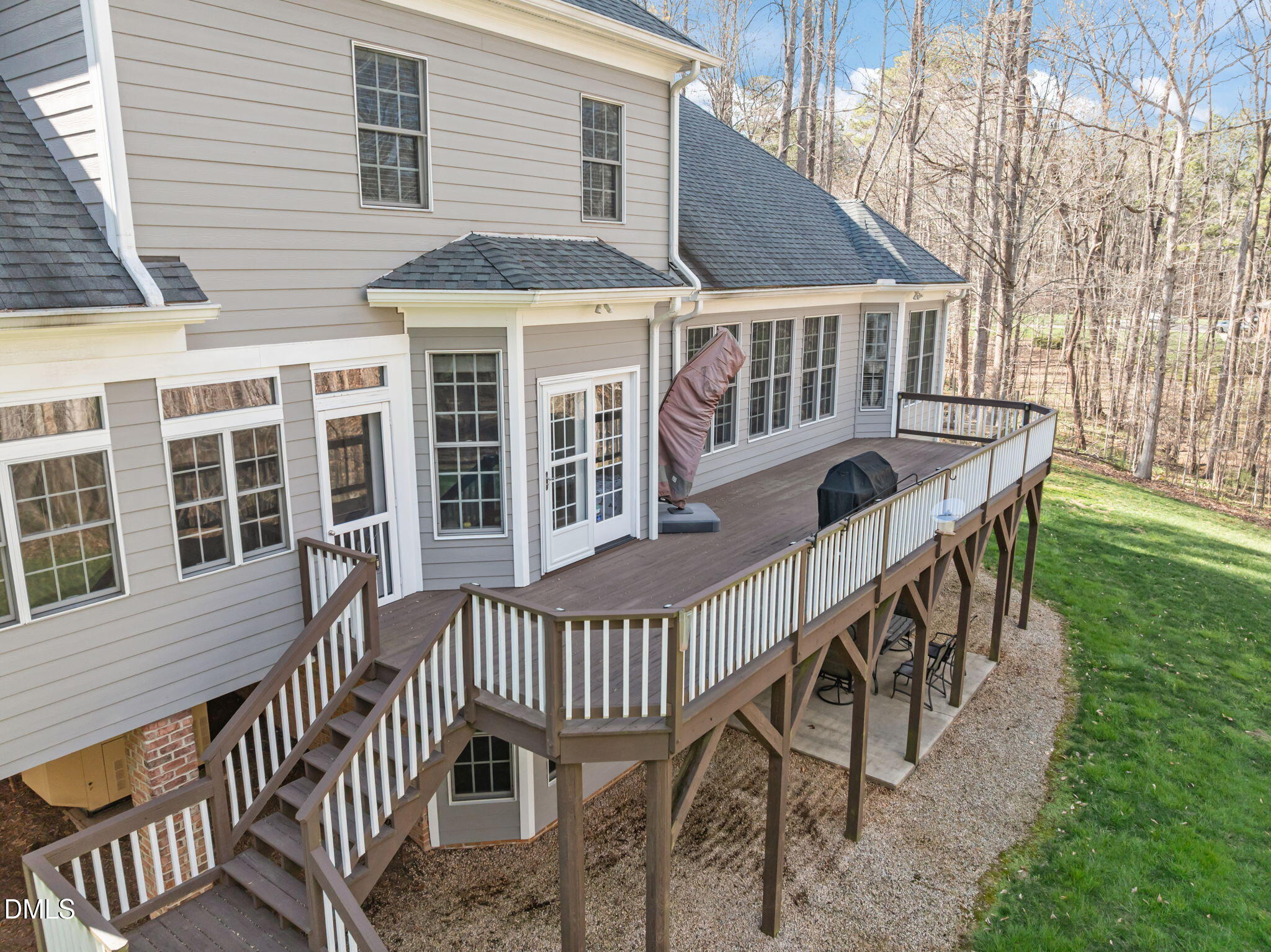House rear with a large elevated deck, white railings, and stairs beside a wooded yard.
