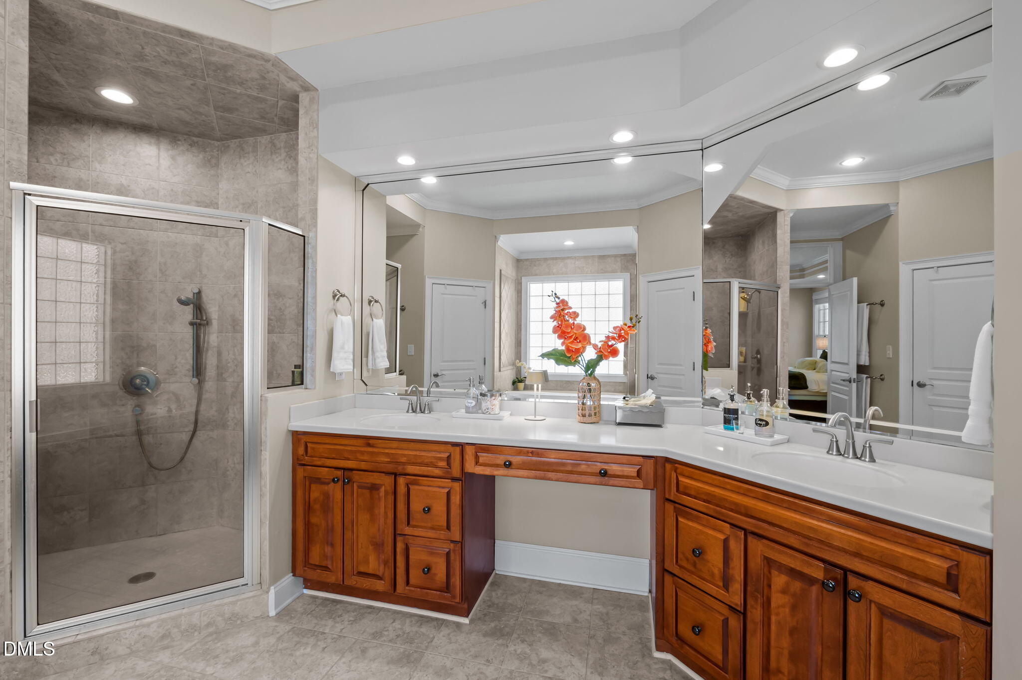 Bathroom with a glass-enclosed tiled shower, long double vanity, and orange flowers by the window.