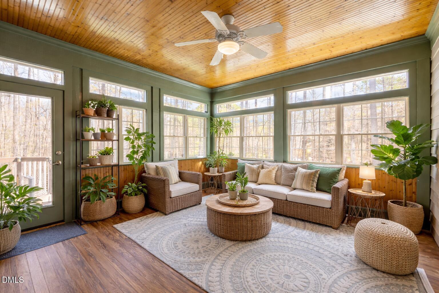 Sunroom with wicker seating, abundant potted plants, and large windows overlooking leafless trees.