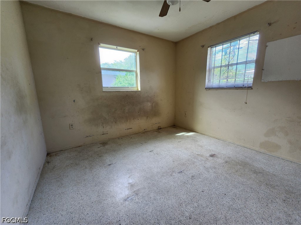 Vacant room with a ceiling fan, two windows, and stained walls and flooring.