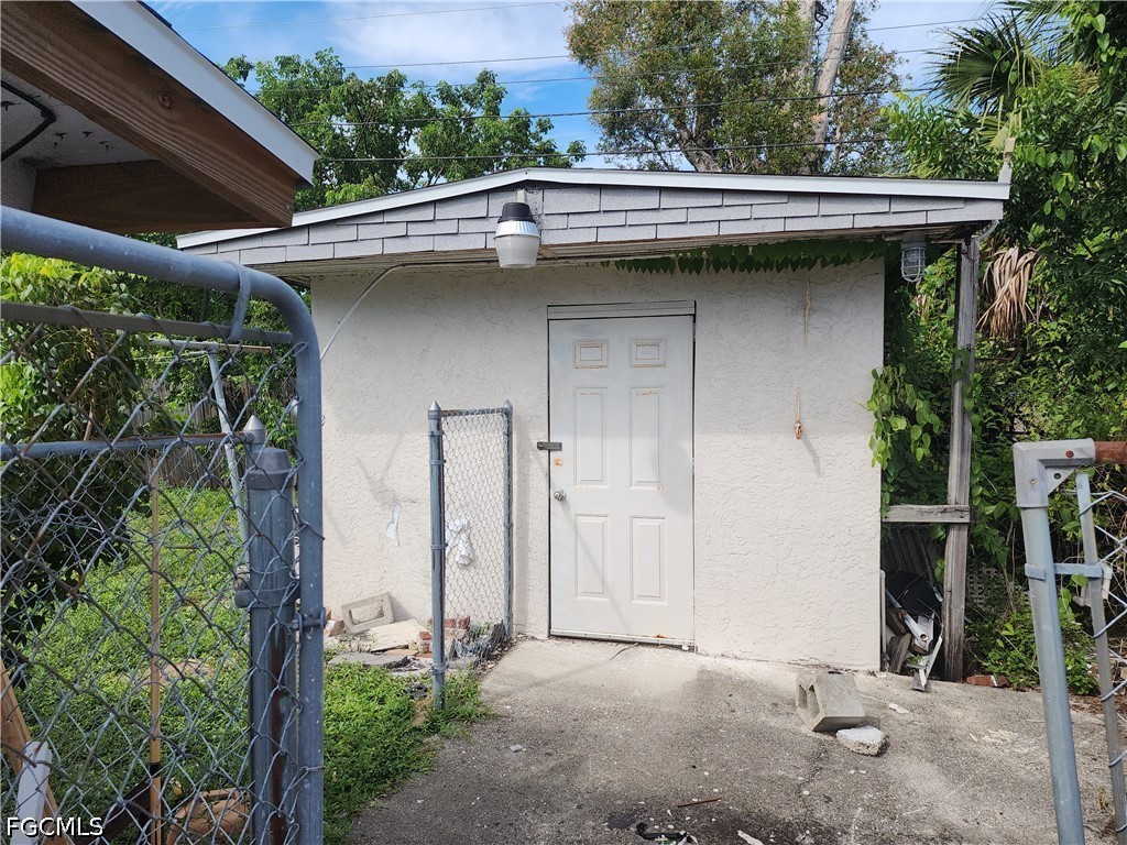 Small white stucco outbuilding with a gray roof, viewed behind a chain-link gate.