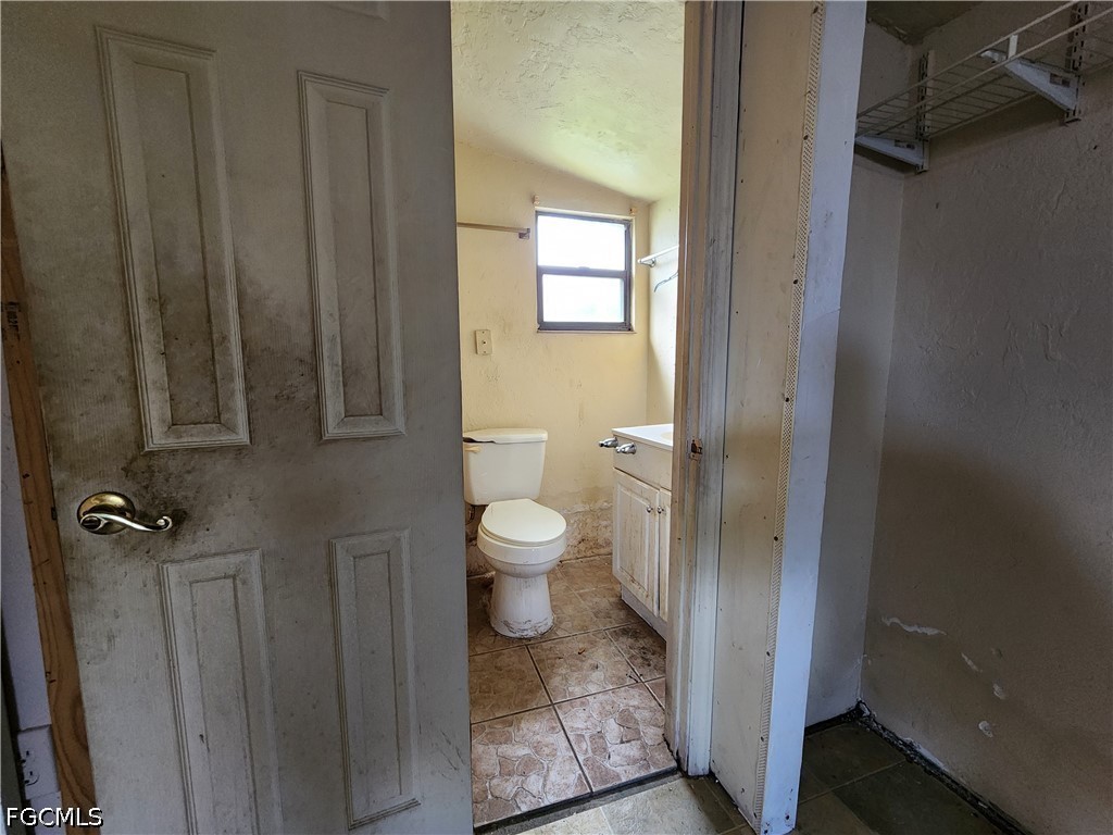 Worn bathroom with a toilet, vanity, tiled floor, and bright window, seen through a partly open door.