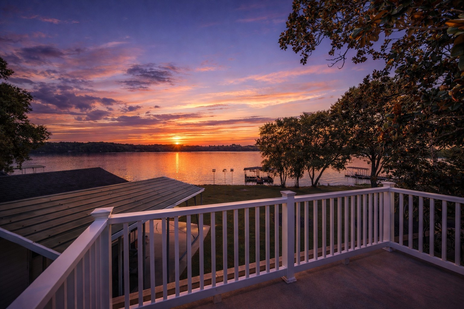 Lake sunset viewed from a deck with white railing and covered docks along the wooded shore.