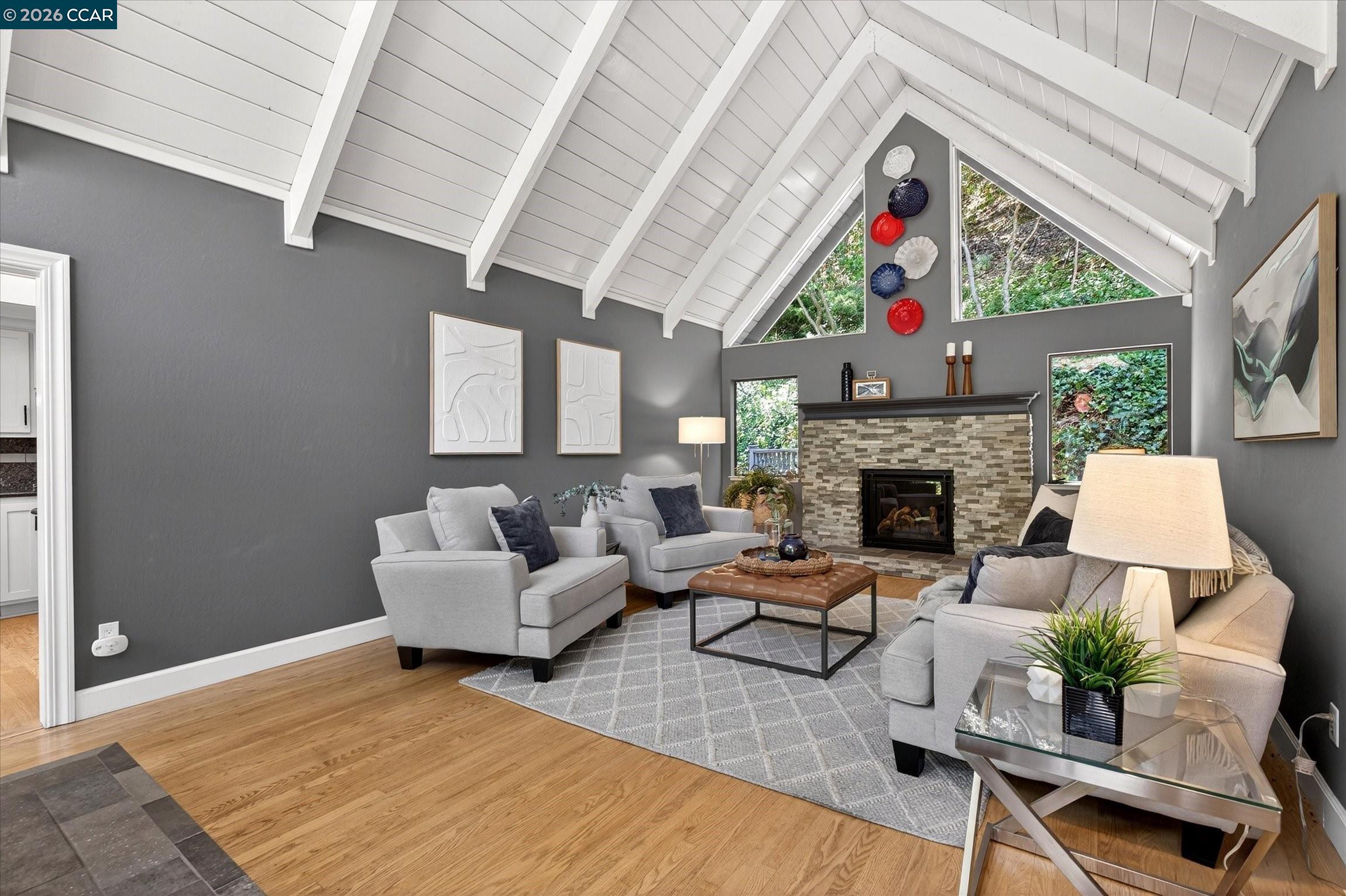 Staged living room with a vaulted beam ceiling, stone fireplace, and triangular windows overlooking trees.