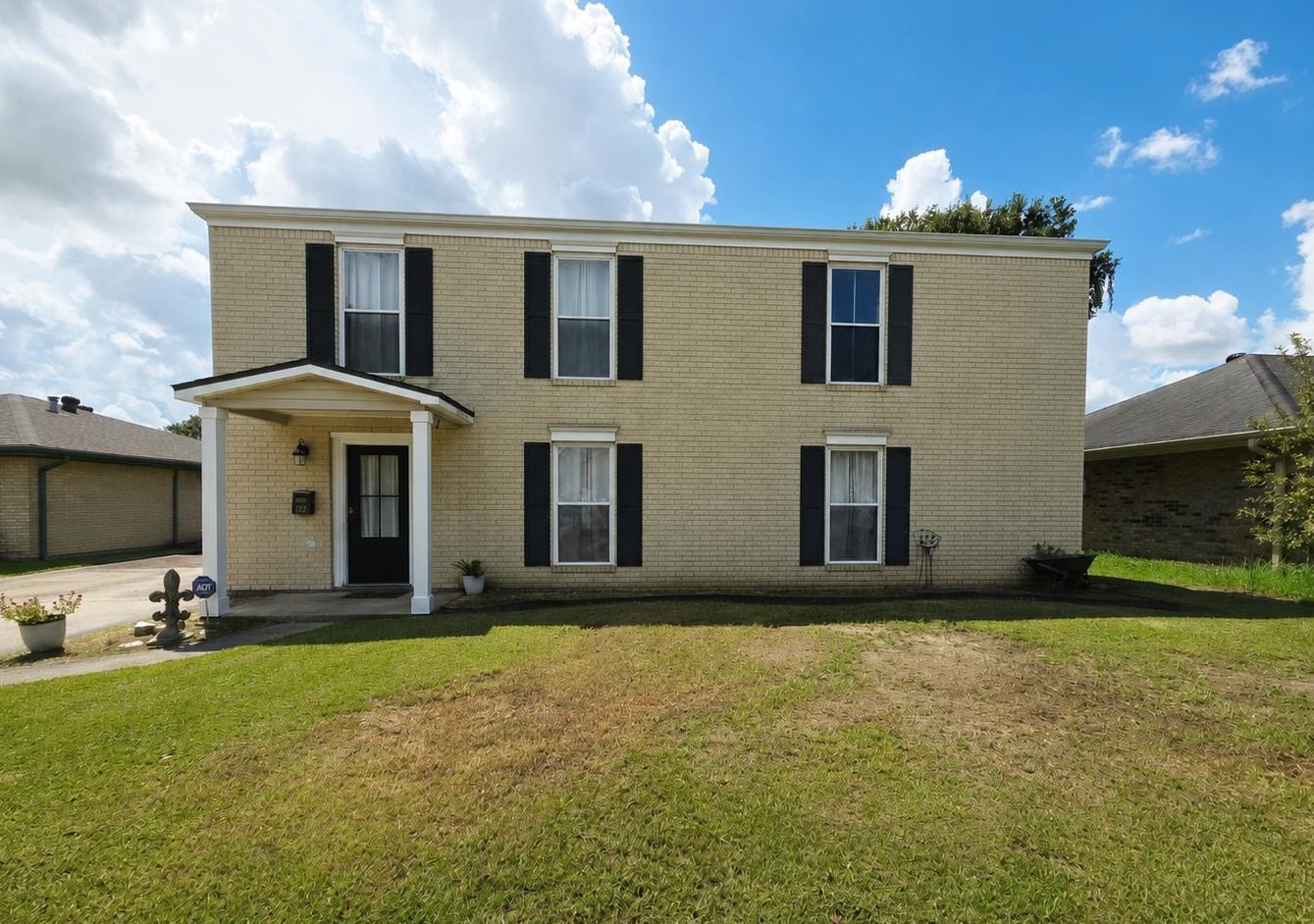 Two-story tan brick house with black shutters, a covered entry, and a broad front lawn.
