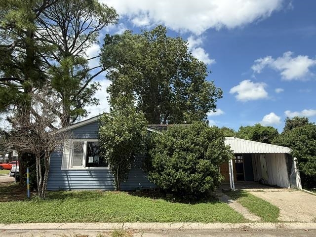 Blue-gray single-story house partly screened by trees, with a covered carport and concrete driveway.