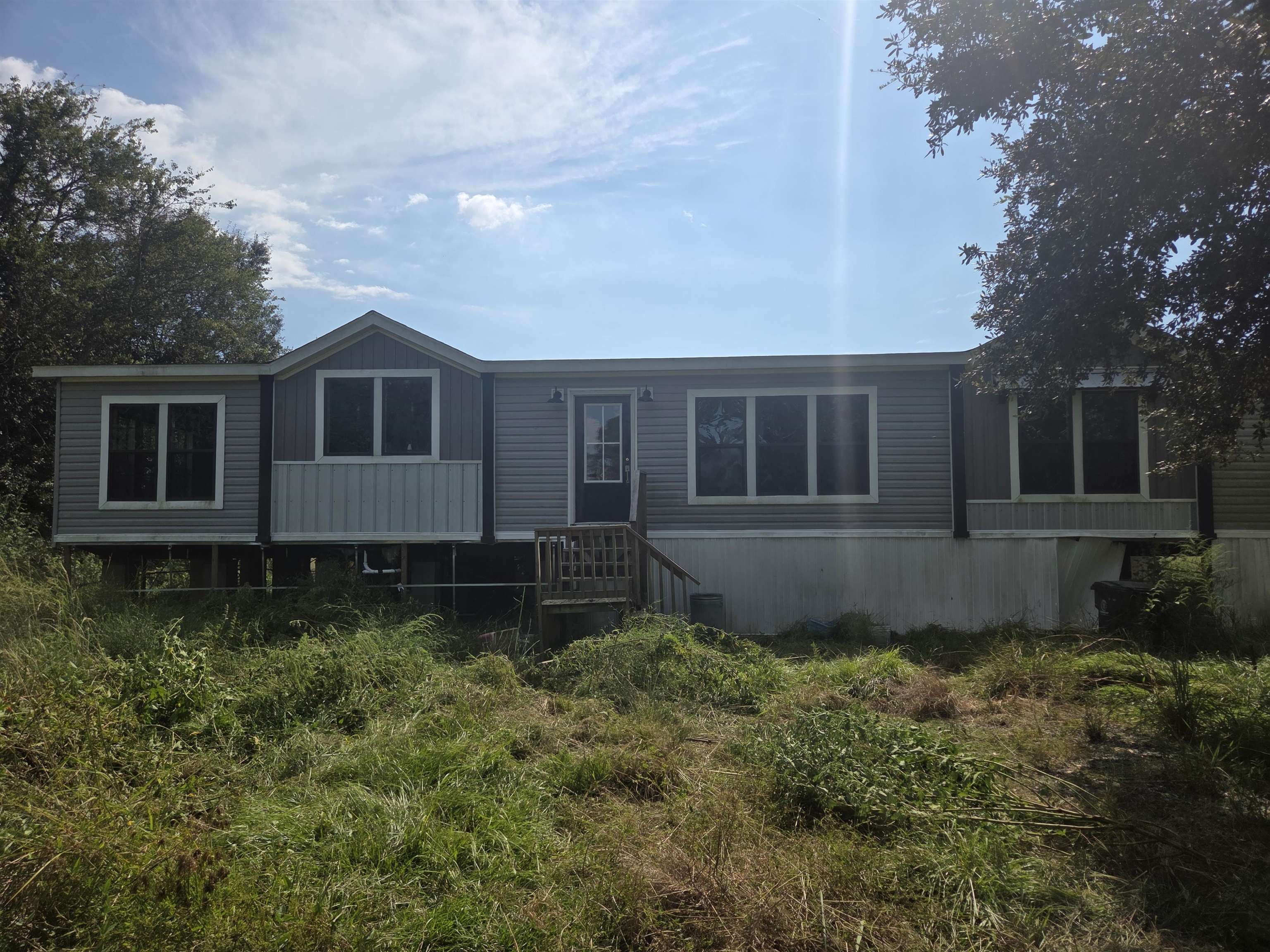 Raised gray home with white trim and a small entry staircase, surrounded by overgrown grass.