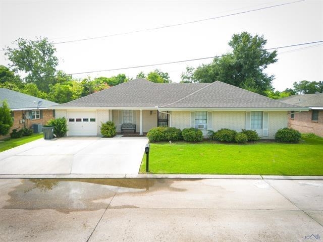Single-story brick house with a gray roof, attached garage, driveway, and bright green front lawn.