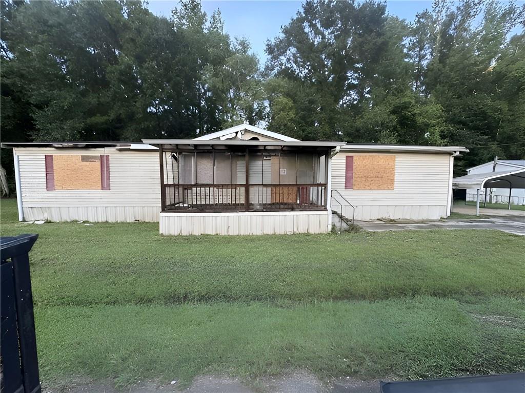 White manufactured home with boarded windows and a covered porch on a grassy, tree-lined lot.