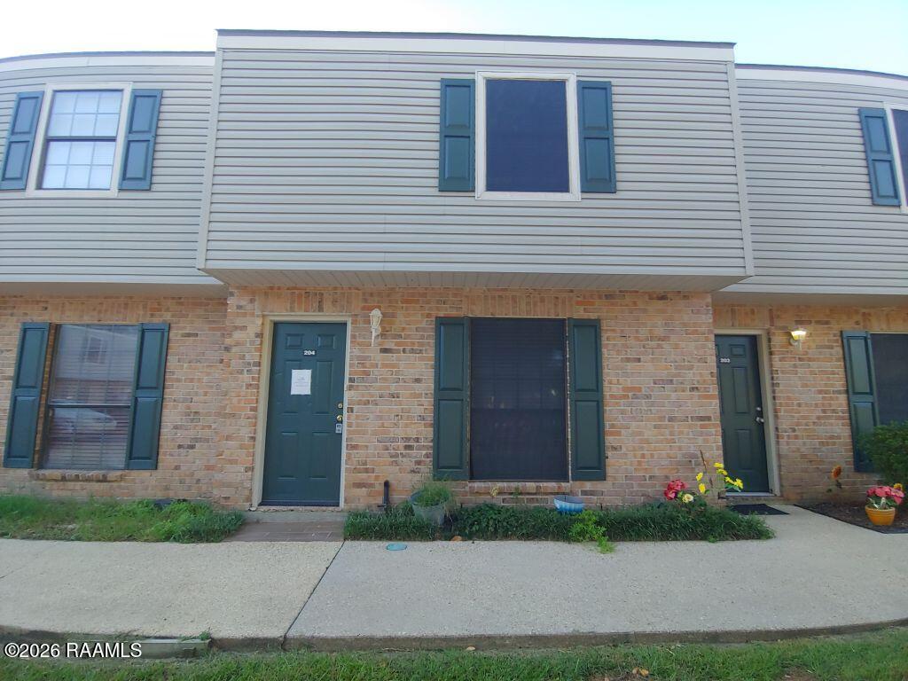 Attached townhouse with tan brick, light siding, teal doors and shutters, and flowers by the sidewalk.