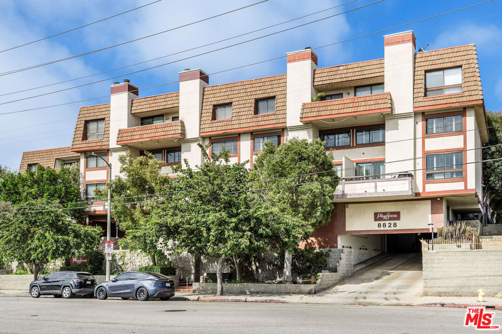 Condominium building with a tiled roof, balconies, trees, and a sloped driveway to the garage.