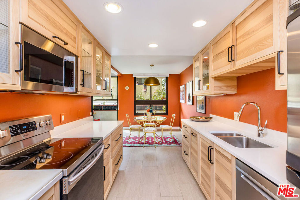 Galley kitchen with light wood cabinets, orange walls, and a dining table by the windows.