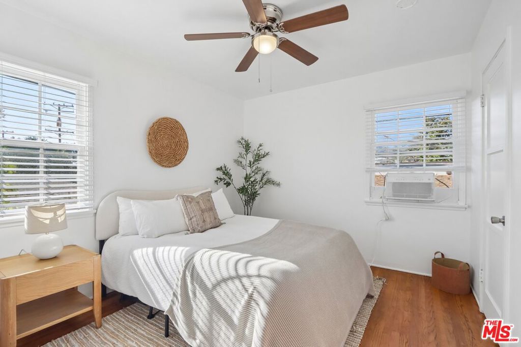 Staged bedroom with a white bed, wood nightstand, ceiling fan, and window air conditioner.