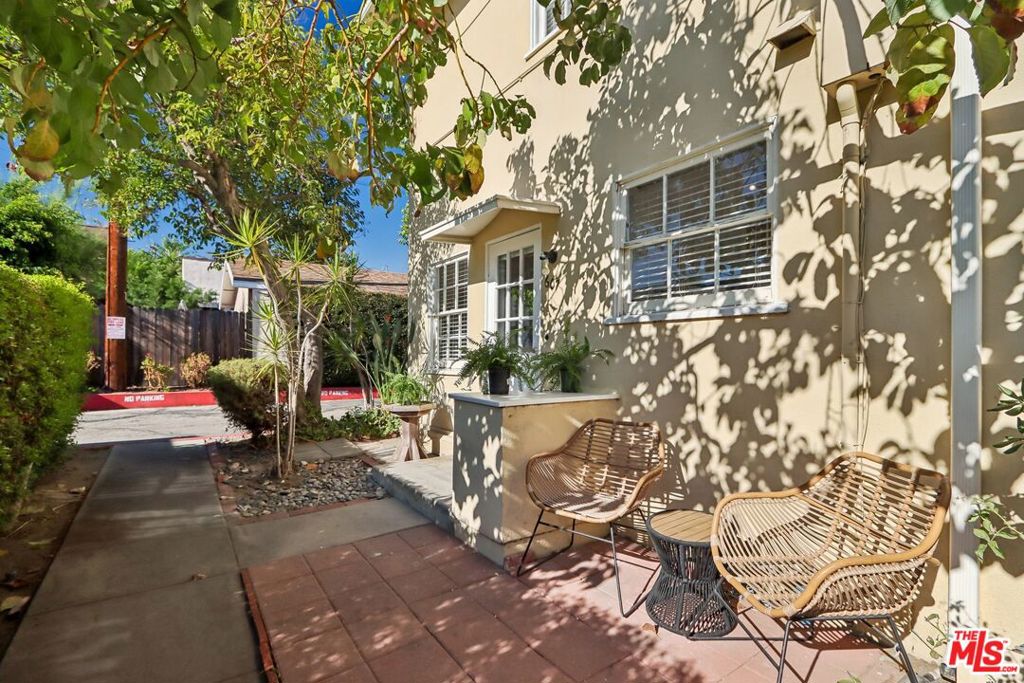 Residential patio with woven chairs and a table beneath leafy trees beside a beige building.