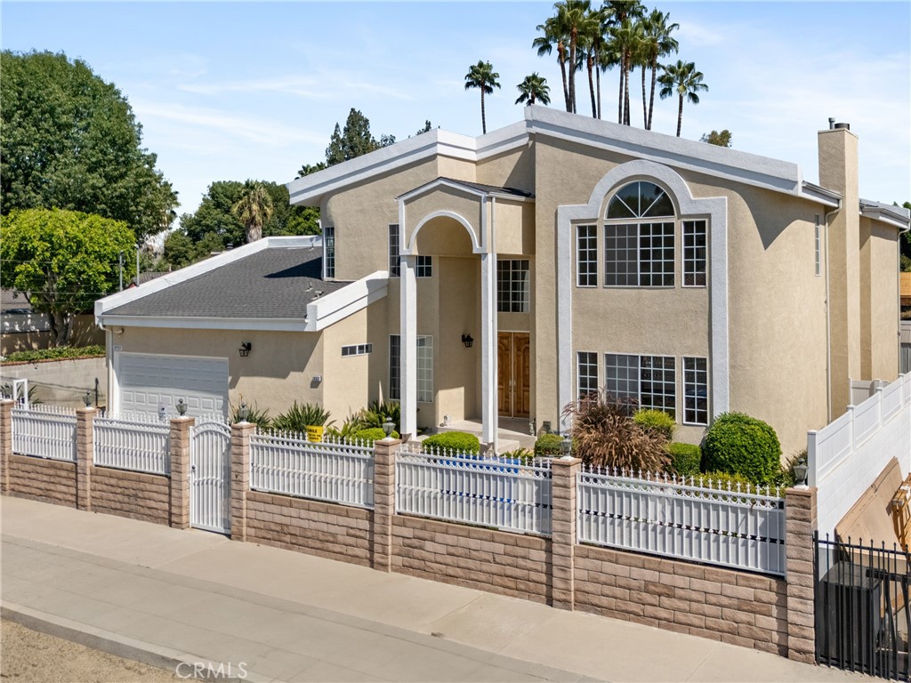 Two-story beige stucco home with arched windows, an attached garage, and a white fence.