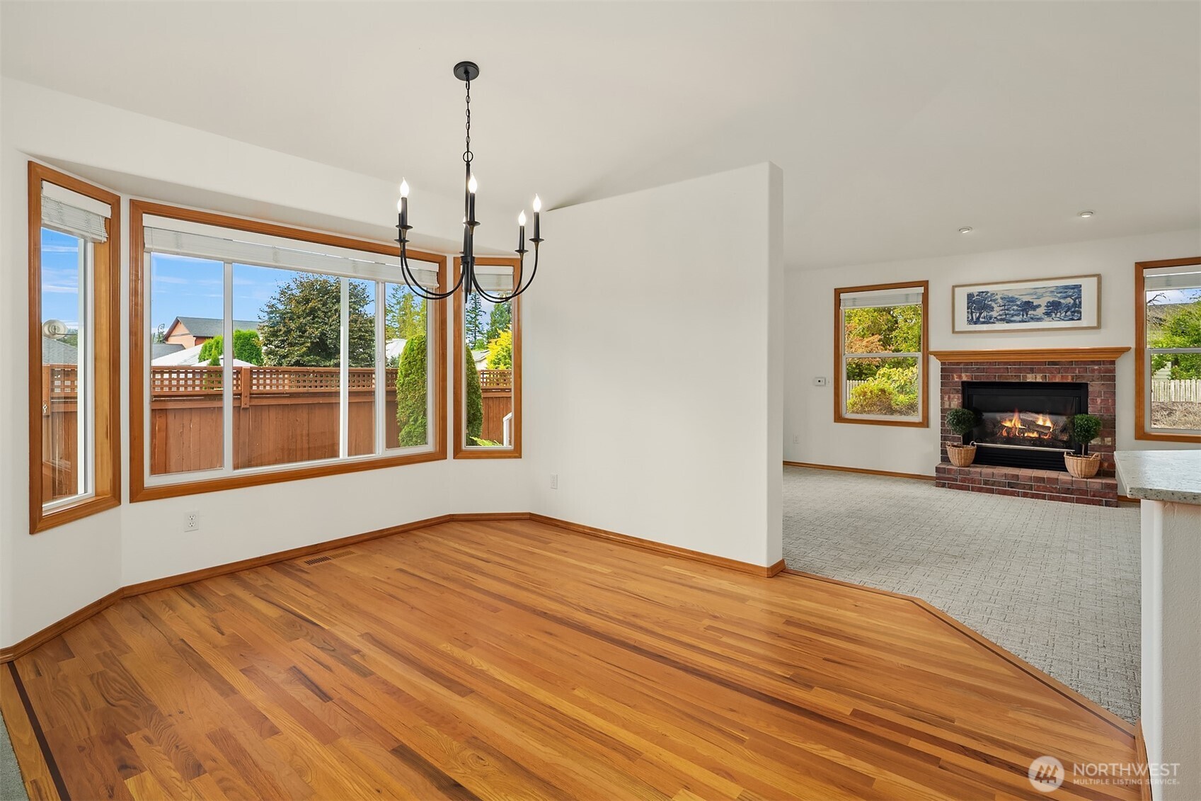 Dining area with hardwood floors and wood-trimmed windows opening to a living room with a brick fireplace.